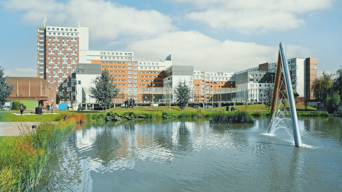An image of Aston University buildings next to a lake with a fountain. 